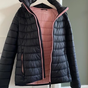 Svart pufferjacka från Everest - Säljer en svart pufferjacka från Everest med rosa foder. Jackan har en dragkedja framtill och en praktisk huva. Perfekt för kyliga dagar med sin quiltade design som håller värmen. Den har långa ärmar och två fickor med dragkedja.