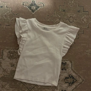 Vit topp från Zara - Säljer en söt vit ribbad topp från Zara med volangdetaljer på ärmarna. Perfekt för en stilren look. Toppen är i en mjuk och bekväm kvalitet. 