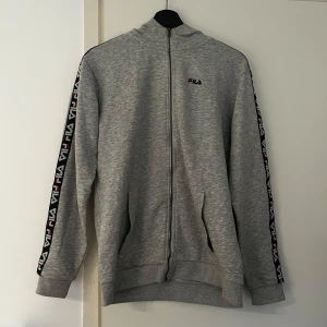 Grå hoodie från FILA - Säljer en grå hoodie från FILA med dragkedja och logotyp längs ärmarna. Perfekt för en sportig look eller avslappnad stil. Den har långa ärmar och en bekväm passform.