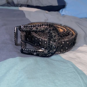 Svart glittrigt bälte med nitar. Sönder på ett ställe med det håller bra endå. - Snyggt svart bälte med glittriga detaljer och nitar. Bältet har en dekorativt utsmyckad spänne och ett korsmönster. Perfekt för att ge din outfit en edgy touch.