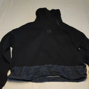 Svart jacka från G-Star RAW - Säljer en svart jacka från G-Star RAW med huva och denimdetaljer vid nederkanten. Jackan har en stilren design med långärmade ärmar och en liten logotyp på framsidan. Perfekt för en casual look.