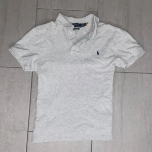 Grå pikétröja från Ralph Lauren - Säljer en vit pikétröja från Ralph Lauren med den ikoniska loggan broderad i blått på bröstet. Tröjan har korta ärmar och en stilren krage med knappar. Perfekt för en avslappnad men stilfull look. Tröjan är i storlek 18-20 junior storlek vilket motsvarar ungefär XS!