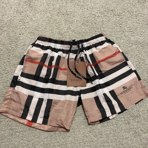 Burberry badshorts - Snygga badshorts från Burberry med klassiskt rutigt mönster i beige, svart, vit och röd. De har en elastisk midja med snörning för perfekt passform. Perfekta för sommarens alla badäventyr! Pris går att diskutera!