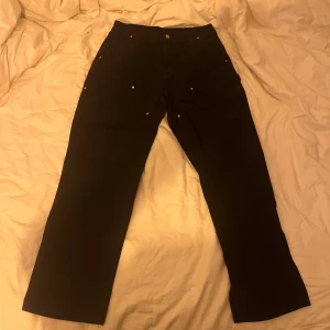 Svarta byxor från Carhartt - Snygga svarta byxor från Carhartt med klassisk design. Byxorna har en rak passform och är perfekta för en avslappnad stil. De har knappar och en liten logotyp på baksidan.