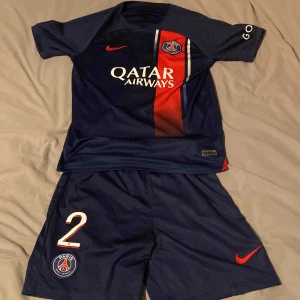 Mörkblå PSG fotbollströja med shorts - Säljer en mörkblå PSG fotbollströja med matchande shorts. Tröjan har korta ärmar och ett rött vertikalt band med klubbens emblem och sponsorlogga. Namnet 'Hakimi' och nummer 2 är tryckt på baksidan. Perfekt för fotbollsfans!