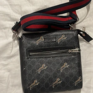 Svart axelväska med tigertryck - Snygg svart axelväska med tigertryck och röd-blå randig axelrem. Väskan har en dragkedja framtill och ett stilrent mönster. Perfekt för att ge din outfit en cool touch.
