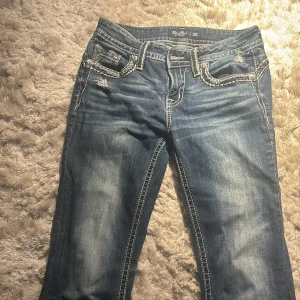 Blå jeans från Miss Me - Snygga blå jeans från Miss Me med coola detaljer och slitningar. De har en lågmidjad passform och dekorativa bakfickor med broderade mönster. Perfekta för en avslappnad stil med en touch av edge.Skriv privat för flera bilder