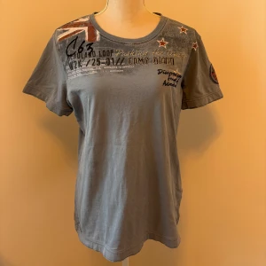 Blå t-shirt från Camp David - Säljer en blå/grå t-shirt från Camp David med tryck och broderade detaljer. T-shirten har ett par fläckar i en lite mörkare färg än resten av tshirten lämgst ut på ena sidan framme.