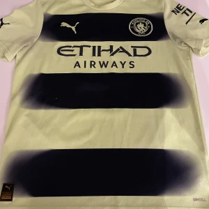 Ljusgul och svart Manchester City fotbollströja från Puma med Haaland på ryggen  - Säljer en snygg Manchester City fotbollströja från Puma i gult med svarta ränder. Tröjan har klubbens emblem på bröstet och Haaland på ryggen. Perfekt för fans av laget! Storlek 15-16 år. 