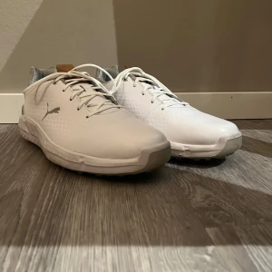 Vita Golfskor från Puma - Snygga vita Golfskor från Puma med diskret logga på sidan. Skorna har snörning och en bekväm sula, perfekt för en stilren look. De har en grå detalj vid hälen och en brun flik på plösen. Använd en gång(pga fel storlek), som nya. Ny pris 1200 kr. Skorna sitter även bra för mig med str 42.