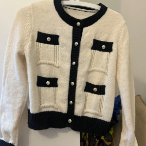 Vit och svart kofta med knappar - Snygg vit kofta med svarta detaljer och knappar. Den har fyra fickor framtill och ribbade kanter. Perfekt för en stilren look.