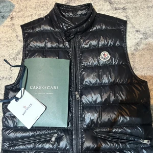 Svart dunväst från Moncler - Säljer en snygg svart dunväst från Moncler med dragkedja och logotyp på bröstet. Västen har en quiltad design och hög krage för extra värme. Perfekt för kyligare dagar.