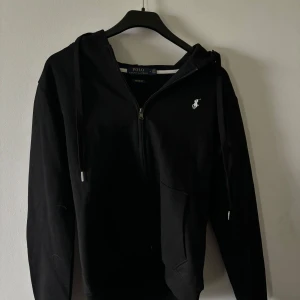 Svart hoodie från Polo Ralph Lauren - Snygg svart hoodie från Polo Ralph Lauren med dragkedja och huva. Den har en broderad logga på bröstet och är i en custom fit. Perfekt för en stilren och avslappnad look. Nästintill oanvänd då den storleken har varit för liten för mig så den är i nyskick