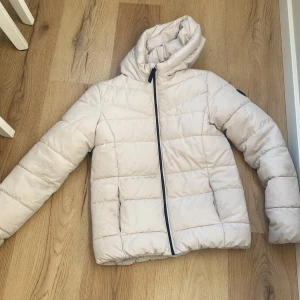 Beige dunjacka med huva - Säljer en snygg beige dunjacka med huva. Jackan har en dragkedja framtill och är perfekt för kyligare dagar. Den quiltade designen ger en stilren look. Passar bra för både höst och vinter.