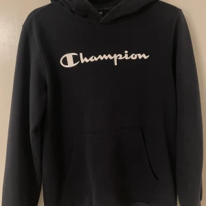 Svart hoodie från Champion - Säljer en svart hoodie från Champion med deras ikoniska logga i vitt på bröstet. Tröjan har en klassisk design med huva och långa ärmar, perfekt för en avslappnad stil. Den är tillverkad i ett mjukt material som ger en bekväm känsla.