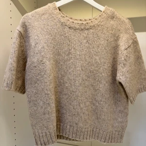 Beige stickad tröja - Säljer en mysig beige stickad tröja med korta ärmar. Perfekt för lager-på-lager under kyligare dagar. Tröjan har en rund halsringning och ribbade kanter vid ärmslut och nederkant.