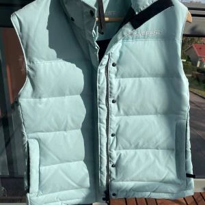 Ljusblå dunväst från Everest - Säljer en snygg ljusblå dunväst från Everest. Västen har en dragkedja framtill och knappar för extra säkerhet. Perfekt för kyliga dagar när du vill hålla värmen utan att kompromissa med stilen. Den är i bra skick och redo för nya äventyr!