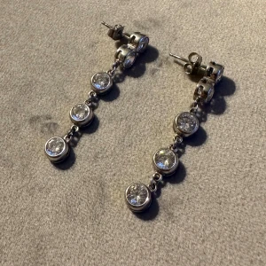 Silverfärgade örhängen med glittrande stenar - Säljer ett par eleganta och stilrena silverfärgade örhängen med flera glittrande stenar i rad. Perfekta för att ge en touch av glamour till vilken outfit som helst. De är långa och hängande, vilket gör dem idealiska för festliga tillfällen.