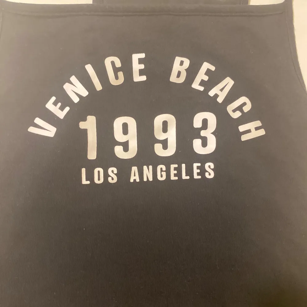 Säljer ett svart linne med tryck 'Venice Beach 1993 Los Angeles' i vitt. Linnet har smala axelband och är perfekt för en avslappnad stil. Passar bra till flares (säljer för jag behöver pengar och varan är knappt använd!). Toppar.