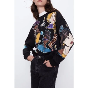 Svart tröja med grafiskt tryck från Zara - Säljer en cool svart tröja från Zara med ett färgglatt grafiskt tryck av olika ansikten och text. Tröjan har långa ärmar och en normal passform. Perfekt för att ge din outfit en unik touch!