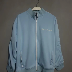 Ljusblå jacka från Palm Angels - Säljer en snygg ljusblå jacka från Palm Angels. Den har en dragkedja framtill och två fickor med dragkedjor. Jackan är långärmad och har en stilren design med Palm Angels-loggan på bröstet. Perfekt för en cool och avslappnad stil!