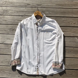 Burberry skjorta - Säljer en stilren vit skjorta från Burberry i storlek XL, men sitter som M-L (är 190 och den passar mig perfekt) Skjortan har långa ärmar och klassiska Burberry-rutiga detaljer på insidan av kragen och ärmsluten. Den är broderad med en diskret logga på bröstet och har knappar framtill.  Äkta men har ej äkthetsbevis, därav priset.