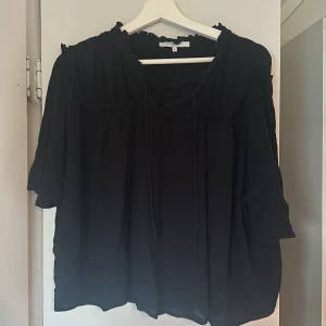 Svart blus med volangdetaljer - Säljer en svart blus med volangdetaljer runt halsen och ärmslut.