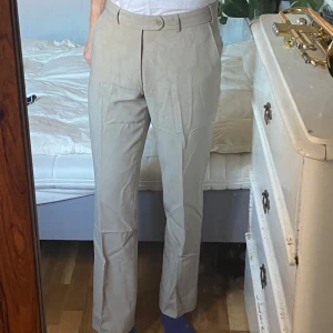 Beige kostymbyxor från Riley - Säljer ett par stilrena beige kostymbyxor från Riley. De har en klassisk design med raka ben och en knapp i midjan. Byxorna är i mycket bra skick och har en snygg passform. Material: 60% ull, 38% polyester, 2% elastan.