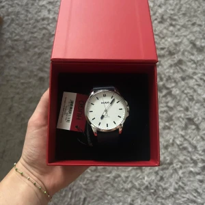 Hugo boss klocka - Säljer en stilren och elegant klocka från Hugo Boss. Den har en klassisk vit urtavla med en silverfärgad boett. Armbandet är i mörkblå skinn, vilket ger en tidlös och sofistikerad look. 
