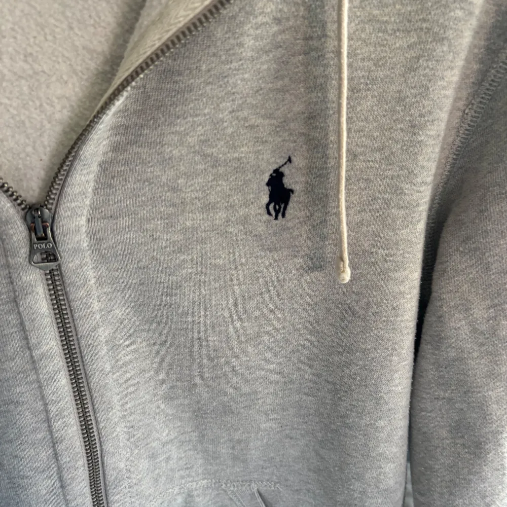 Säljer en snygg och bekväm grå hoodie från Ralph Lauren. Bra skick lite nopprig som man kan se på sista bilderna. Den har en dragkedja framtill och en klassisk logga broderad på bröstet. Perfekt för höst och vår, med en mysig huva och långa ärmar. Passar både till vardags och chillkvällar. 🩶. Tröjor & Koftor.