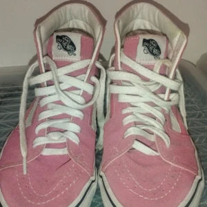 Rosa och vita sneakers från Vans - Säljer ett par snygga rosa och vita Vans sneakers i storlek dam 39. De har en klassisk hög modell med snörning och den ikoniska vita sidoremsan. Skorna är i bra skick och perfekta för både vardag och fest. Passar till alla dina outfits!
