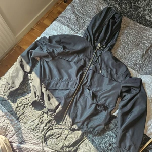 Svart vindjacka från H&M - Säljer en svart vindjacka från H&M i storlek S. Jackan har en dragkedja framtill och en justerbar huva med dragsko. Perfekt för blåsiga dagar! Den är i bra skick och har elastiska muddar vid ärmsluten för extra komfort.