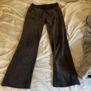 Mörkgråa mjukisbyxor med snörning - Säljer ett par mörkgråa mjukisbyxor från Juicy Couture. De har en avslappnad passform med snörning i midjan för justerbarhet. Byxorna har fickor både fram och bak med knappar på bakfickorna. Tyvärr så har en av metallklämmorna ramlat av.