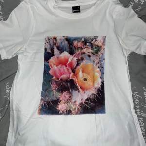 Säljer en snygg vit t-shirt från Gina Tricot i storlek S. Den har ett färgglatt blommigt tryck på framsidan med rosa och gula blommor. Perfekt för en avslappnad stil eller att liva upp en enkel outfit. T-shirten är i bomull och har korta ärmar.