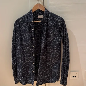 Mönstrad skjorta från Knowledge Cotton Apparel - Säljer en snygg mörkblå skjorta från Knowledge Cotton Apparel i storlek M. Skjortan har ett unikt vitt mönster och knappar framtill. Perfekt för både vardag och fest!