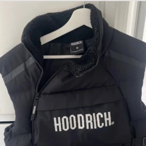 Svart dunväst från Hoodrich - Säljer en svart dunväst från Hoodrich i mycket bra skick. Västen har en stor ficka framtill med dragkedja och justerbara remmar på sidorna. Perfekt för höst och vinter, både stilren och praktisk. Passar bra för en casual look eller streetwear-stil.