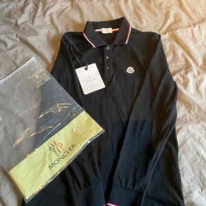 Svart pikétröja från Moncler - Säljer en snygg svart pikétröja från Moncler i nytt skick. Tröjan har långa ärmar och klassisk krage med röd och vit rand. Perfekt för både vardag och fest! 
