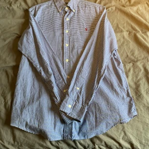 Blårutig skjorta från Ralph Lauren - Säljer en snygg blårutig skjorta från Ralph Lauren i storlek M. Skjortan har långa ärmar och klassiska knappar framtill. Den är broderad med Ralph Laurens ikoniska logga i rött på bröstet. Perfekt för både vardag och fest! 💙
