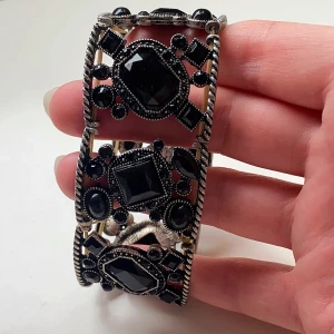 Svart och silver armband med stora stenar - Snyggt och iögonfallande armband i silverfärg med stora svarta stenar. Armbandet har en bred design med detaljerade mönster och en elastisk passform som gör det enkelt att ta på och av. Perfekt för att ge en elegant touch till din festoutfit!