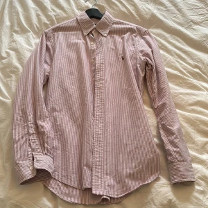 Randig skjorta från Ralph Lauren - Säljer en klassisk långärmad skjorta från Ralph Lauren i vitt och rött randigt mönster. Skjortan har en broderad logga på bröstet och knappar framtill. Perfekt för både vardag och fest! 🕺storlek medium slim fit