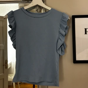 Blå ärmlös topp med volangdetaljer - Säljer en superfin blå ärmlös topp med volangdetaljer längs ärmhålen. Toppen är ribbad. Passar både till vardags och fest! 💙
