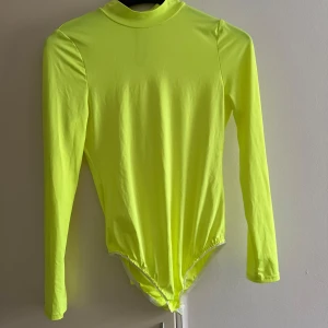 Neongul långärmad bodysuit - Säljer en supercool neongul bodysuit med långa ärmar. Perfekt för att sticka ut på festen eller vardagar. Den har en hög krage och är gjord i ett stretchigt material som sitter tight och bekvämt. Passar perfekt till både vardag och speciella tillfällen!