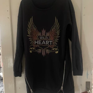 Svart tröja med tryck från Gina Tricot - Cool svart tröja från Gina Tricot med 'Wild Heart' tryck och vingar i guld och rött. Den har trekvartsärmar och dragkedjor på sidorna för extra edge. Perfekt för en avslappnad men stilren look. 🖤✨
