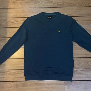 Blå tröja från Lyle & Scott - Säljer en snygg blå tröja från Lyle & Scott i storlek L. Tröjan är tillverkad av 84% bomull och 16% polyester, vilket gör den både bekväm och hållbar. Den har en klassisk design med ett litet gult emblem på bröstet. Perfekt för både vardag och fest!