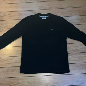 Svart långärmad tröja från Tommy Jeans - Säljer en klassisk svart långärmad tröja från Tommy Jeans i storlek M. Tröjan är i bomull och polyester, perfekt för både vardag och fest. Den har en liten logga på bröstet och är i fint skick. Passar perfekt till höst och vinter! 🍂
