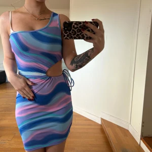 Flerfärgad oneshoulder klänning - Säljer en superfin oneshoulder klänning i blå, rosa och lila toner. Den har en cool cut-out detalj på sidan och knytning i midjan. Perfekt för sommarens fester eller en utekväll. Materialet är stretchigt och bekvämt. Passar perfekt för dig som vill sticka ut! Prislappen kvar!