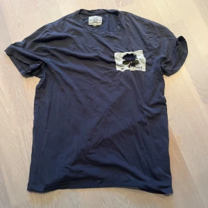 Mörkblå t-shirt från Kent & Curwen - Säljer en mörkblå t-shirt från Kent & Curwen i bomull. Den har en cool patch på bröstet med ett tryck och årtalet 1926. Perfekt för en avslappnad stil. T-shirten är kortärmad och passar bra till både jeans och shorts.