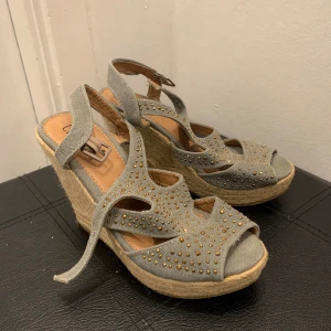 Grå kilklackssandaler med strass - Snygga grå sandaler med kilklack och öppen tå. De har dekorativa strassdetaljer och en bekväm rem runt ankeln. Perfekta för sommarens fester eller en dag på stan. Klacken är flätad i naturmaterial vilket ger en bohemisk touch.