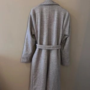 Grå kappa från 157 - Säljer en stilren grå kappa från 157 i storlek M. Den har ett snyggt bälte i midjan och långa ärmar. Perfekt för höst och vår! Kappan är i mycket bra skick och har en klassisk design som passar till det mesta. 🧥✨