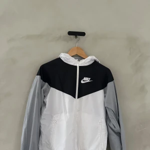 Svart och vit vindjacka från Nike - Säljer en snygg svart och vit vindjacka från Nike. Perfekt för vår och höst med sin lätta design och dragkedja framtill. Jackan har en huva och långa ärmar, vilket gör den idealisk för blåsiga dagar. Den har också praktiska fickor med dragkedja. Passar både till träning och vardagsbruk.